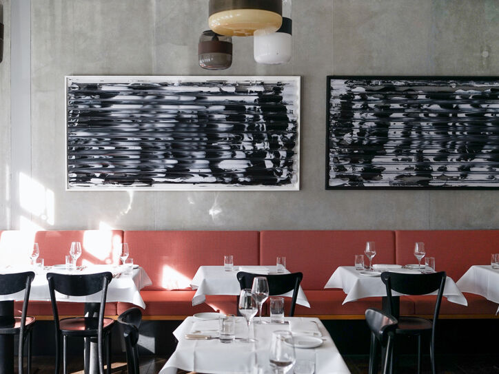 Ein lichtdurchfluteter Raum mit mehreren eingedeckten Tischen, weißen Tischdecken, schwarzen Stühlen, einer rot-braunen durchgängigen Sitzbank und zwei großen Bildern mit abstrakten Schwarz-Weiß-Motiven.