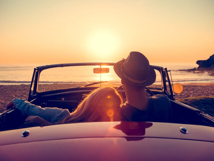 Rückansicht eines Paares in einem Cabriolet an einem Sandstrand bei Sonnenuntergang, sie hat den Kopf auf seine Schulter gelehnt.
