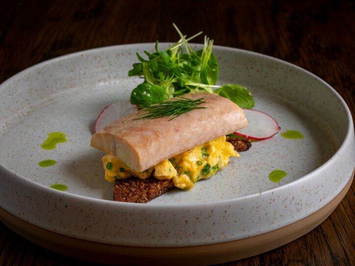 Ein Keramikteller mit hohem Rand, auf dem ein Stück Fisch mit weiteren Zutaten hübsch angerichtet liegen.