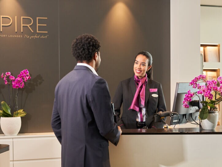 Eine lächelnde Frau in Uniform begrüßt einen Kunden am Empfangstresen einer Flughafenlounge.
