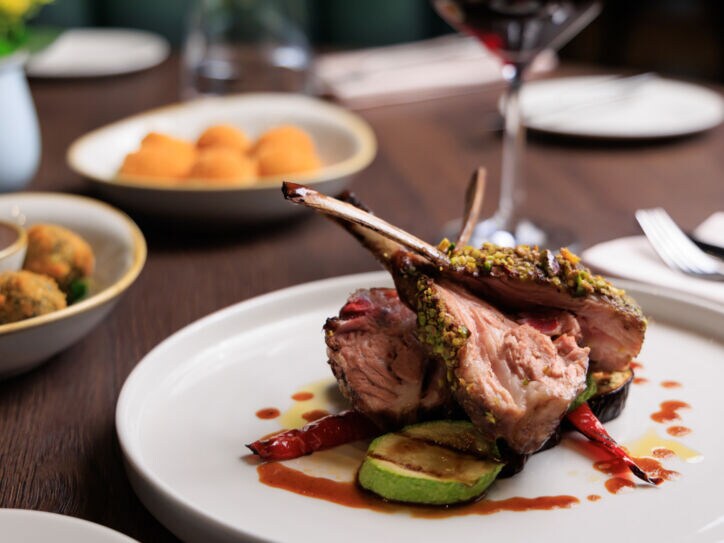 Gegrilltes Lammrippenkotelett, angerichtet auf einem weißen Teller, daneben Beilagen auf einem Tisch in einem Restaurant.