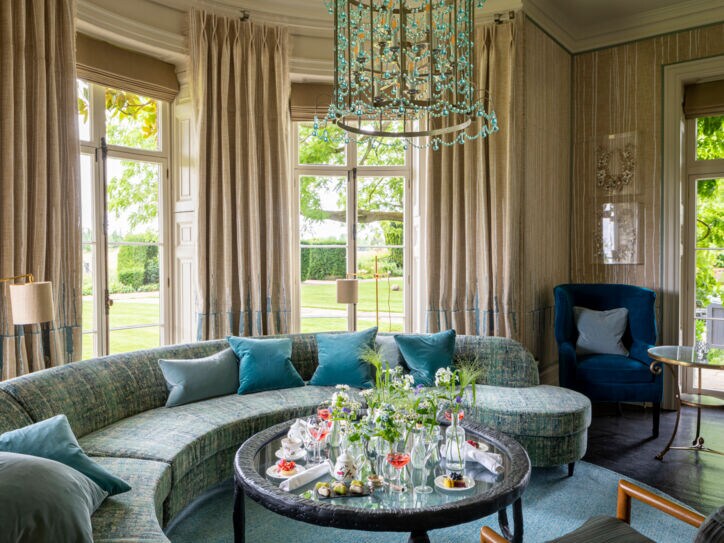 Luxuriöser Salon mit Kronleuchter und geschwungenem Sofa vor Panoramafenstern mit Blick in einen Garten.