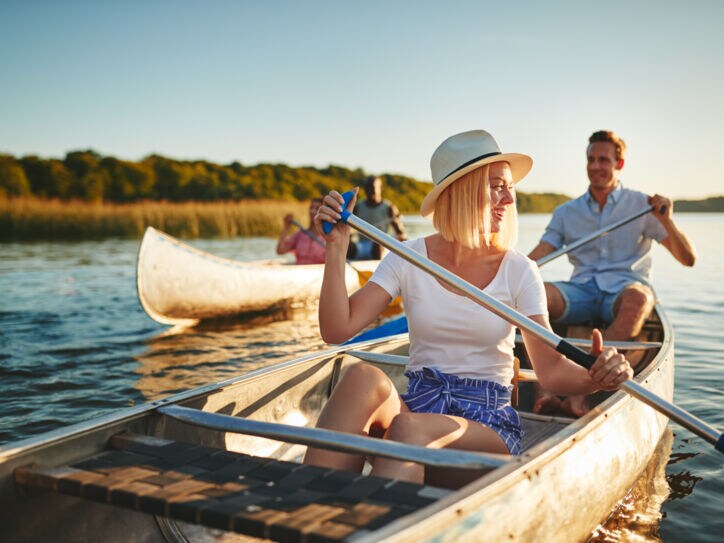 Zwei Paare in sommerlicher Kleidung rudern in Kanus auf einem Gewässer bei abendlichem Sonnenschein.