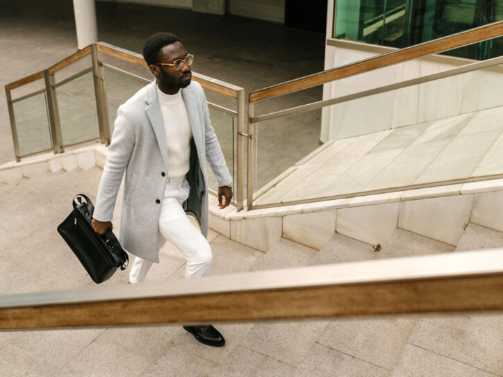 Ein elegant gekleideter Mann in einem hellen Outfit mit Wollmantel und schwarzer Aktentasche läuft eine Außentreppe hoch.