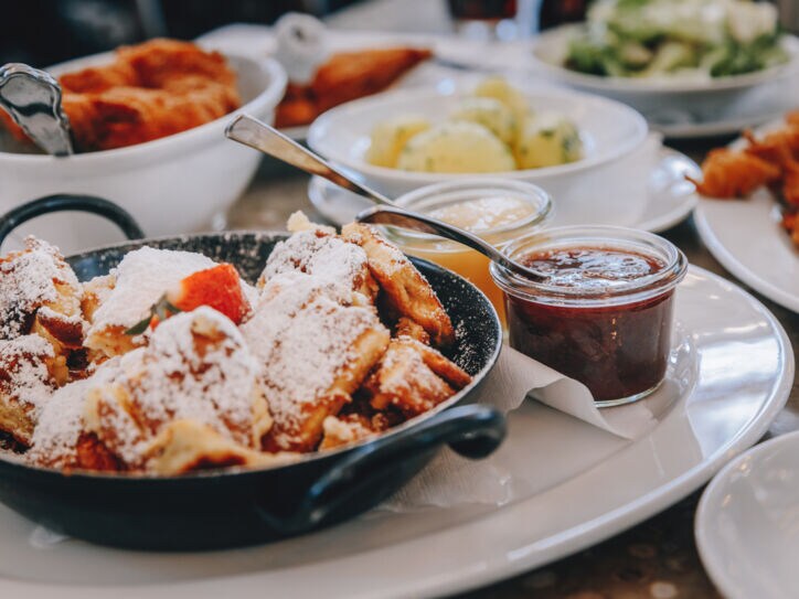 Ein schwarzer Topf mit Kaiserschmarrn auf einem weißen Teller, drumherum kleine Schälchen mit verschiedenen Leckereien.