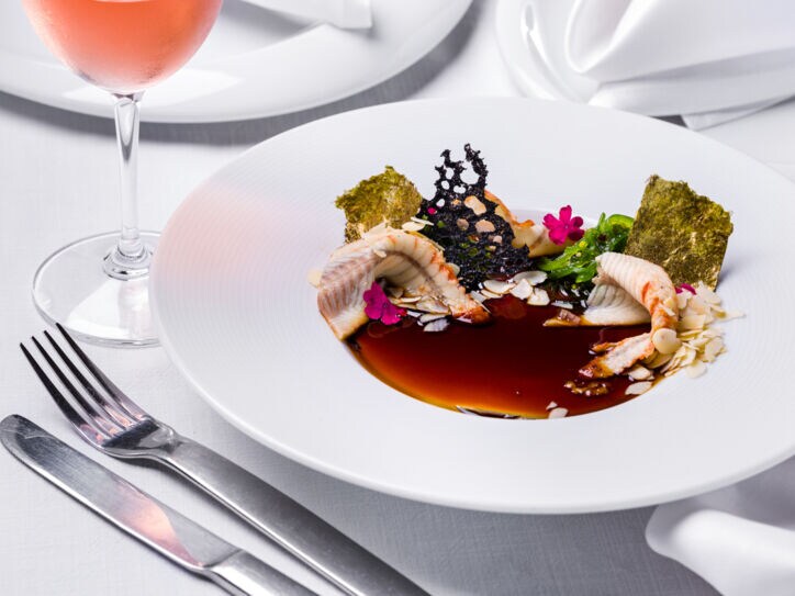 Ein kreativ angerichtetes Fischgericht auf einem weißen Teller auf einem weiß eingedeckten Tisch.