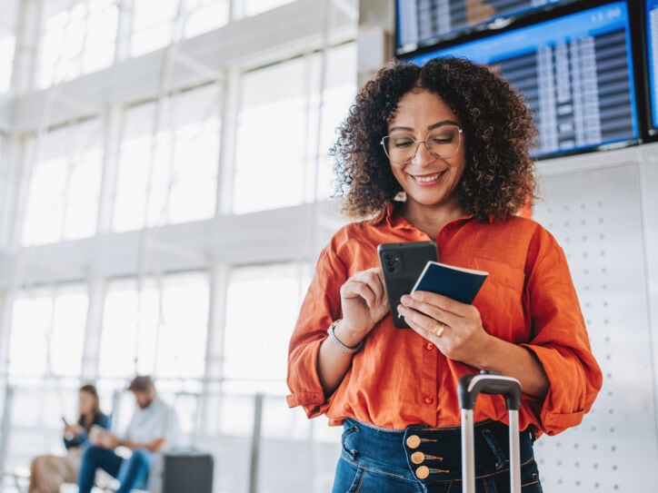 ine Frau bedient lächelnd ihr Smartphone in einem Flughafenterminal, in ihrer Hand ein Reisepass.