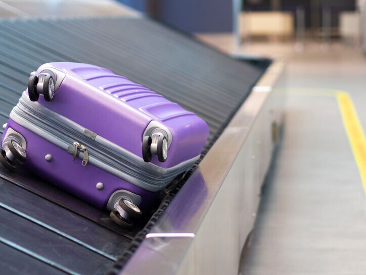 Ein einzelner, lilafarbener Hartschalenkoffer auf einem Gepäckband am Flughafen.