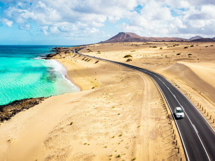 Ein weißes Auto fährt auf einer Schnellstraße durch eine Wüstenlandschaft am türkisblauen Meer.