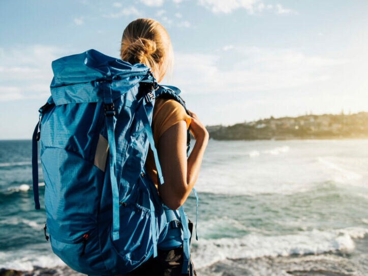 Rückansicht einer Frau mit großem blauen Rucksack am Strand, die aufs Meer blickt.