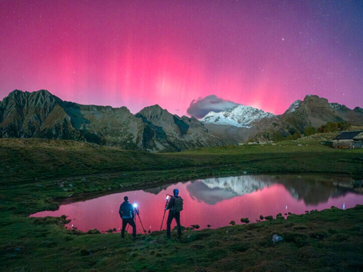 Zwei Menschen stehen vor einer Bergkulisse und fotografieren Polarlichter, die den Himmel pink färben.