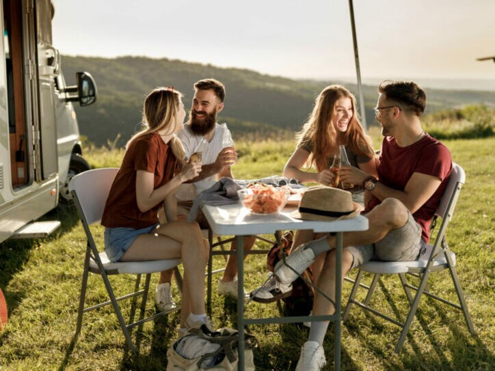Vier gut gelaunte Personen in Sommerkleidung sitzen an einem Campingtisch neben einem Wohnmobil auf einer Rasenfläche vor Hügellandschaft.