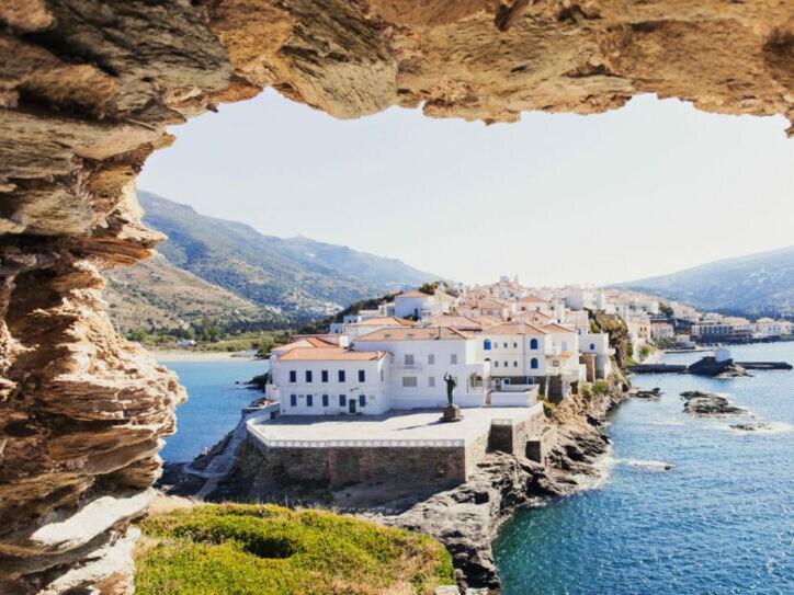 Blick auf weiße Häuser eines Ortes auf einer felsigen Landzunge im Meer durch ein Loch in einer Felsenmauer.