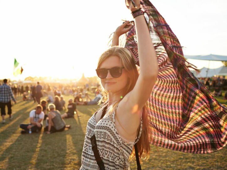 Eine Frau mit Sonnenbrille steht auf einer Wiese, auf der Leute sitzen, und hält ein Tuch über ihren Kopf.