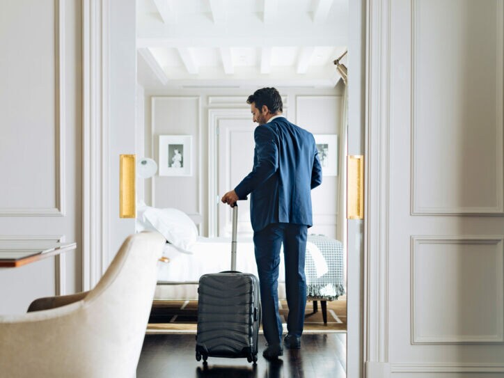 Mann im Anzug und einem Rollkoffer in der Hand betritt eine helle luxuriöse Hotel-Suite.