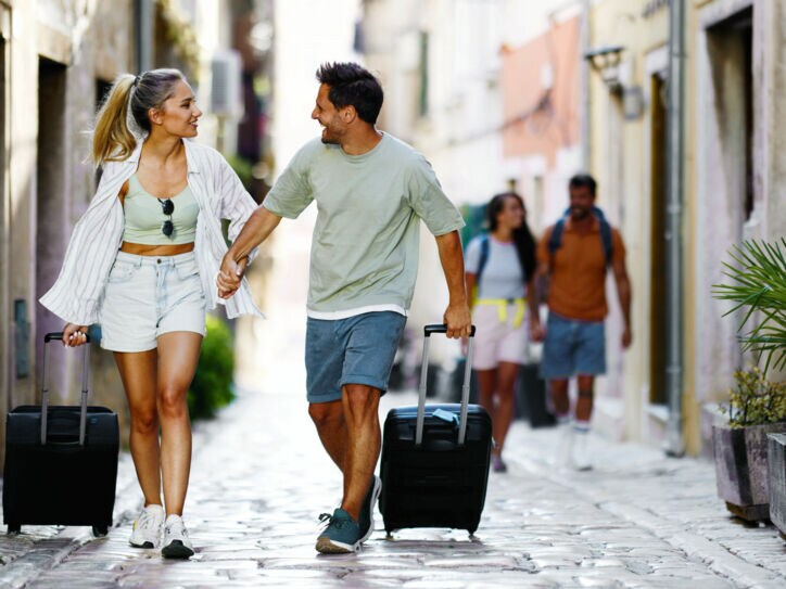 Ein händchenhaltendes Paar in Sommerkleidung geht mit Rollkoffern durch eine Gasse mit Kopfsteinpflaster.
