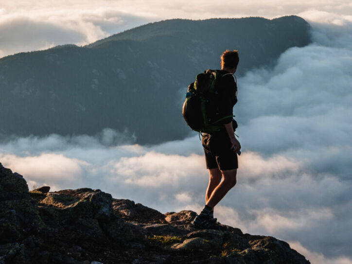 Wanderer mit Rucksack steht auf felsigem Gipfel und blickt auf nebelverhangene Berge bei Sonnenuntergang