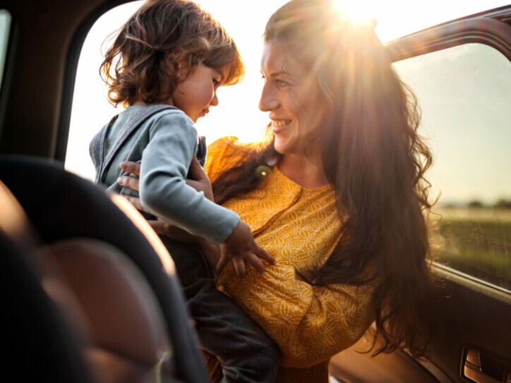 Frau in gelbem Kleid hält lächelnd ein kleines Kind im Auto bei Sonnenuntergang