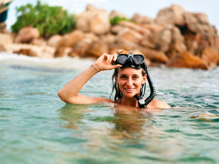 Frau mit Schnorchelmaske im Wasser vor felsiger Küste, hebt die Maske lächelnd an