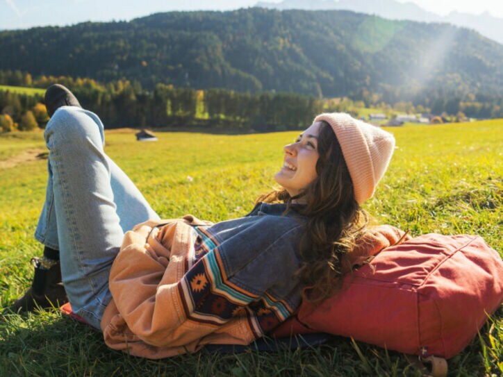 Eine Frau ruht sich auf einer Wiese in der Sonne aus, dabei liegt sie auf ihrem Wanderrucksack.