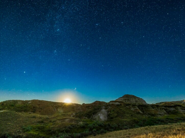 Klarer, tiefblauer Sternenhimmel mit aufgehendem Mond über einer felsigen Graslandschaft.