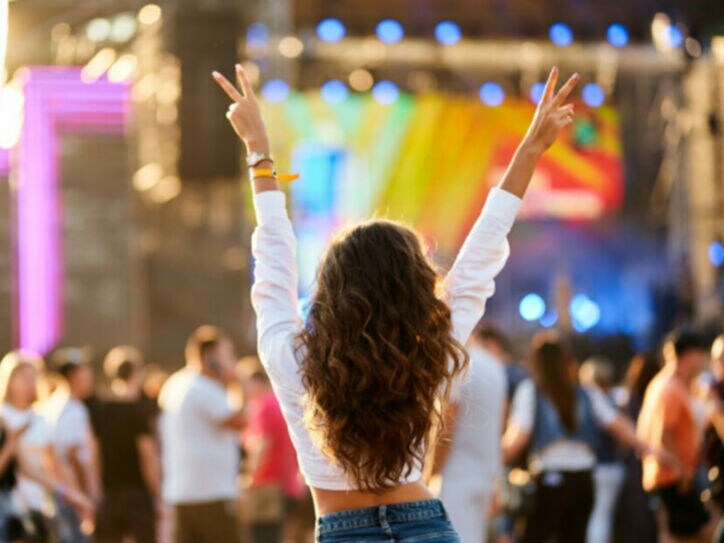 Rückansicht einer jungen Frau, die ihr Hände mit Peace-Zeichen in die Luft hebt vor einer Musikbühne mit Publikum im Freien.