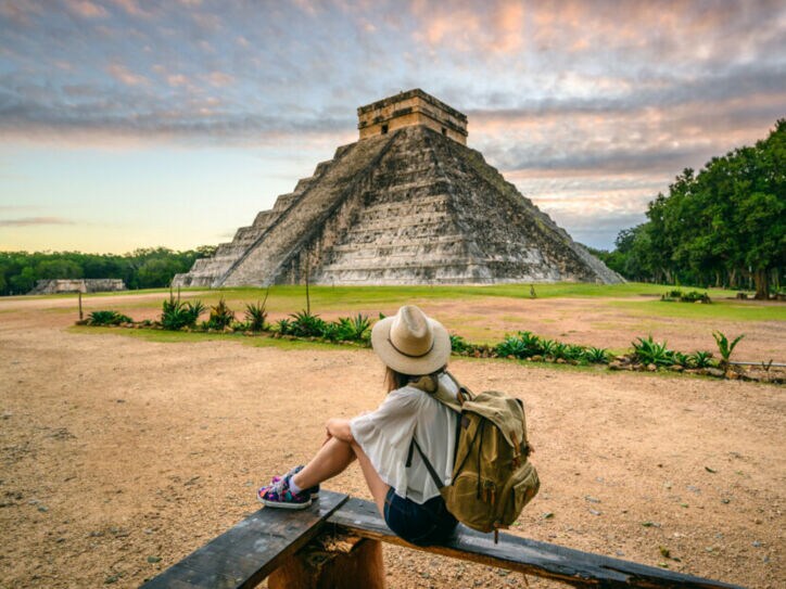 Person mit Hut und Rucksack sitzt auf einer Bank und blickt auf die Pyramide von Chichén Itzá bei Sonnenuntergang