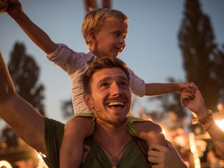 Mann im Dunkeln mit kleinem Kind auf den Schultern, im Hintergrund sind Bäume und Beleuchtung zu sehen.