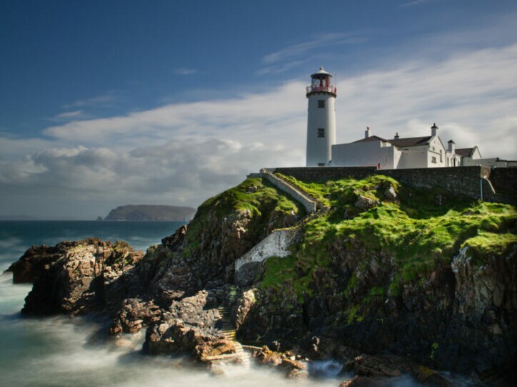 Ein Leuchtturm mit rotem Licht auf einem Hügel, umgeben von grünem Gras und steiniger Küste, mit Wellen, die gegen die Felsen schlagen.
