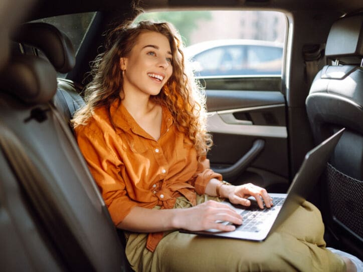 Eine lachende Frau in orangefarbener Bluse sitzt auf dem Rücksitz eines Autos mit einem aufgeklappten Laptop auf ihrem Schoß.
