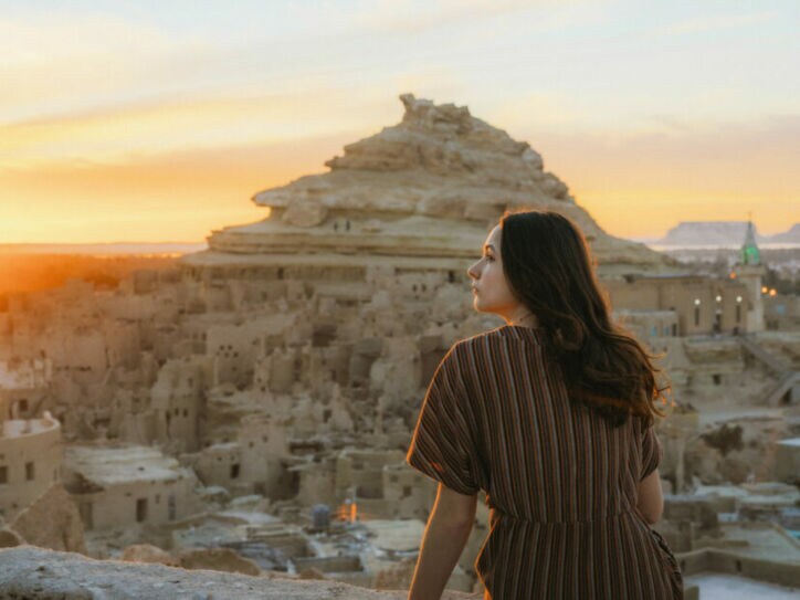 Frau mit braunem gestreiftem Kleid sitzt auf einer Mauer und blickt auf die Felsenstadt in der Siwa-Oase bei Sonnenuntergang in Ägypten.