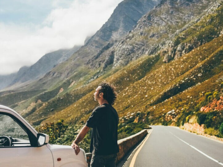 Mann lehnt an weißem Auto an einer Bergstraße, umgeben von steilen Felsen und bewölktem Himmel.