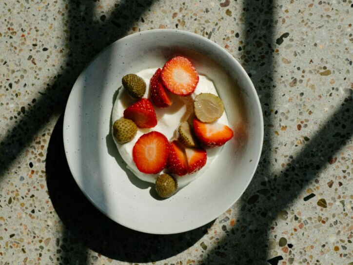 Schale mit Joghurt, belegt mit geschnittenen Erdbeeren und kleinen grünen Früchten, auf marmoriertem Untergrund mit Schatten.