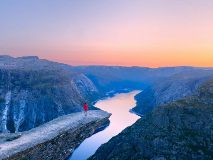 Bergklippe Trolltunga mit Person in roter Jacke, die auf dem Felsvorsprung über einem tiefen Fjord steht, umgeben von Bergen im Abendlicht.