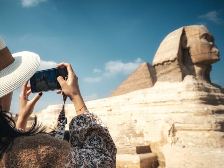 Eine Frau mit Hut fotografiert die Große Sphinx von Gizeh mit einem Smartphone bei klarem Himmel.