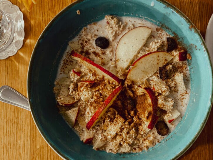 Blaue Schüssel mit Porridge, garniert mit Apfelscheiben, Blaubeeren und Zimt, auf Holztisch mit Glas Wasser und Serviette.