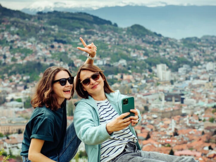 Zwei junge Frauen sitzen auf einer Mauer mit Blick auf eine Stadt, eine macht ein Selfie mit einem Smartphone und zeigt ein Peace-Zeichen.