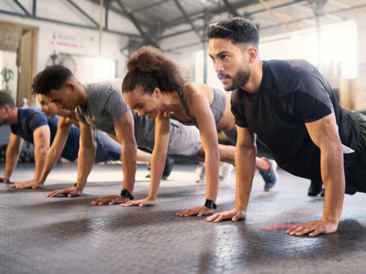 Mehrere Personen in Sportkleidung machen Liegestütze in einem hellen Fitnessstudio mit Gummiboden.