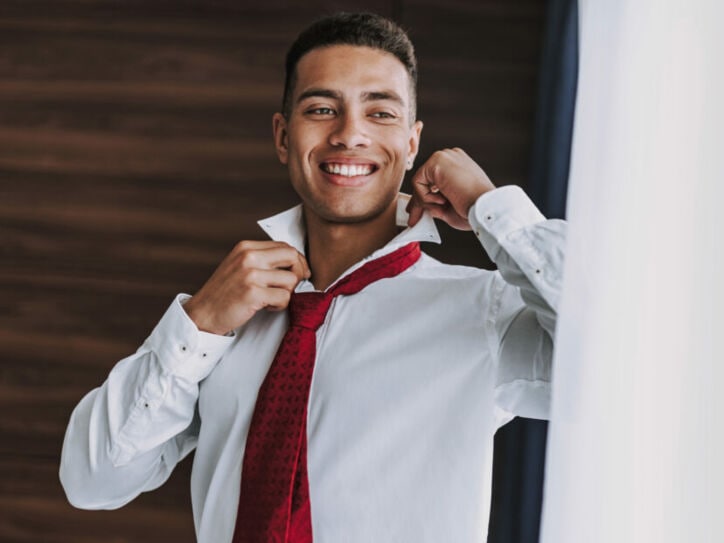 Ein junger Mann im weißen Hemd mit roter Krawatte richtet sich lächelnd den Kragen vor einem Spiegel.