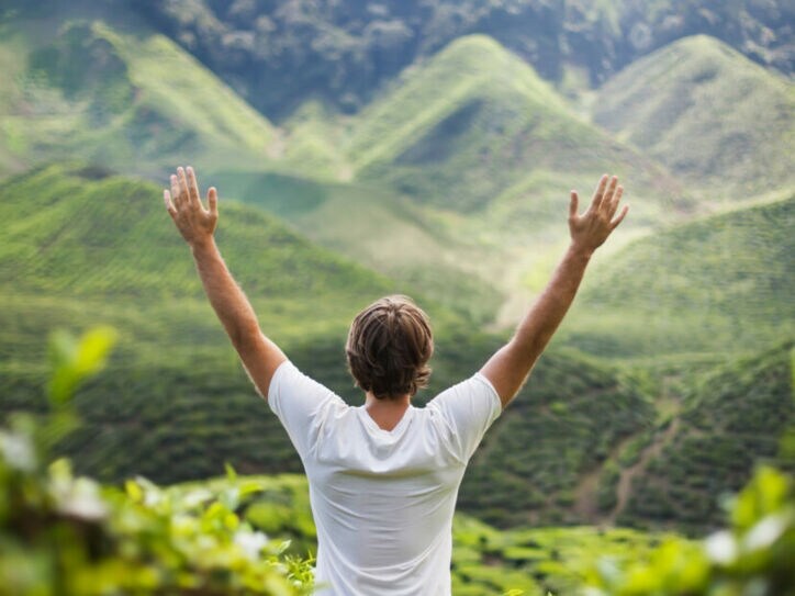 Mann mit weißen T-Shirt steht mit erhobenen Armen vor grünen, bewaldeten Hügeln.