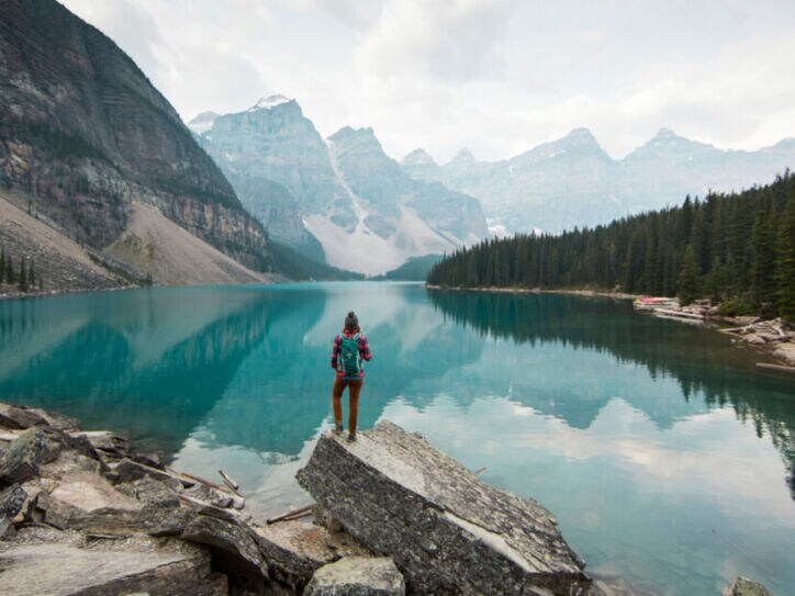 Person mit rotem Rucksack steht auf Felsen am Ufer eines klaren Bergsees, umgeben von Nadelwald und hohen Bergen.