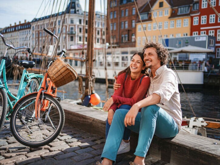 Ein lachendes Paar sitzt eng beieinander auf einer Kaimauer an einem Kanal mit bunten Häusern im Stadtzentrum bei Sonnenschein, neben ihnen stehen zwei Fahrräder auf Kopfsteinpflaster.