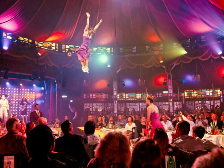 Personen sitzen beim Dinner an Tischen in einem Spiegelzelt während einer Show mit Akrobatinnen.
