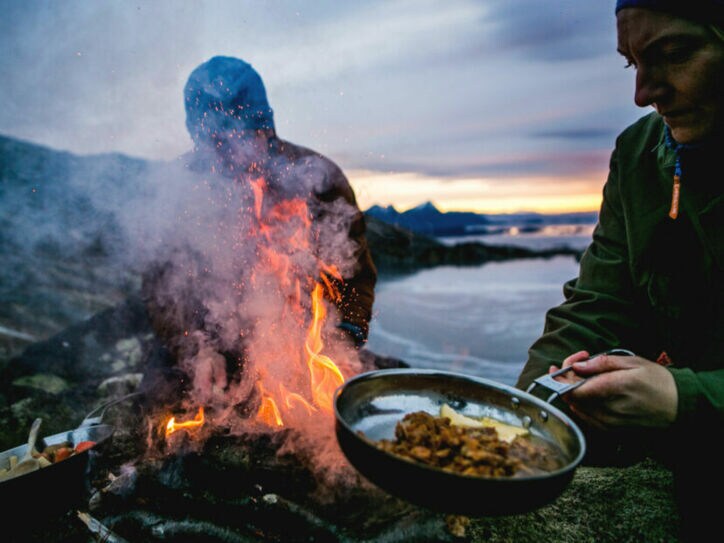Zwei Personen bereiten Speisen in Pfannen über einem Feuer an einer schroffen Felsküste zu bei Abenddämmerung.