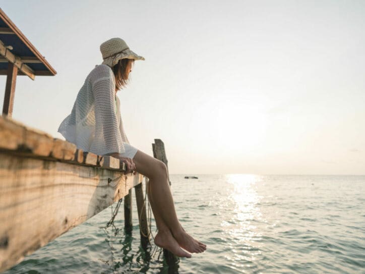 Frau mit Sonnenhut und barfuß, die auf einem Holzsteg sitzt und aufs Wasser schaut.