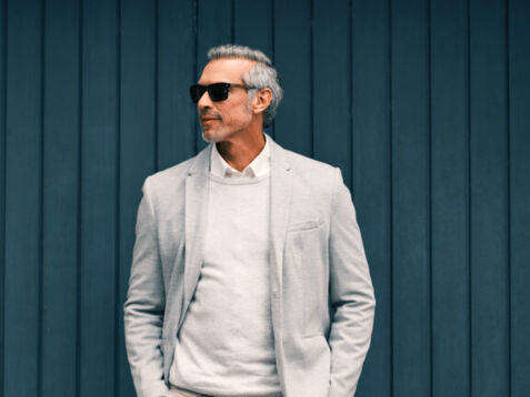 Mann vor einer blauen Holzwand mit weißem Pullover und hellem Anzug mit Sonnenbrille