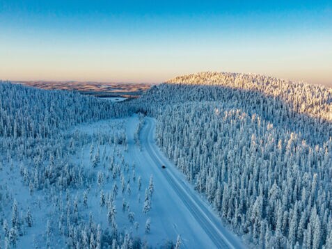 Luftaufnahme einer weiten Winterlandschaft mit schneebedeckten Wäldern, durch die ein einzelnes Auto über eine Straße durch den Schnee in der Abenddämmerung fährt