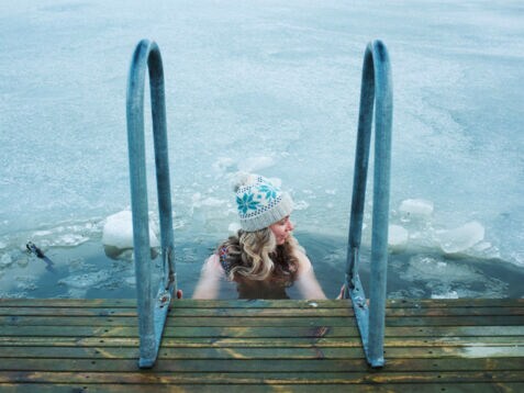 Eine Frau mit Wollmütze hält sich beim Baden in einem zugefrorenen See an dem Handlauf einer Treppe an einem Holzsteg fest.
