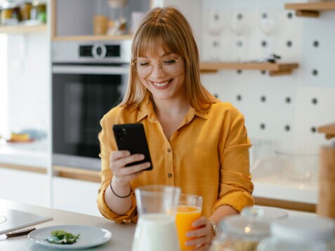 Eine junge Frau in gelbem Hemd steht mit einem Glas Orangensaft in der Hand in der Küche und schaut lächelnd auf ihr Smartphone.