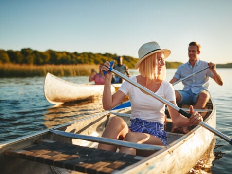 Zwei Paare in sommerlicher Kleidung rudern in Kanus auf einem Gewässer bei abendlichem Sonnenschein.